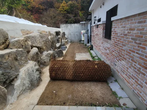 돌담과 벽돌 외벽 사이 흙바닥에 코이어 매트와 멀칭롤이 놓인 마당 시공 준비 현장