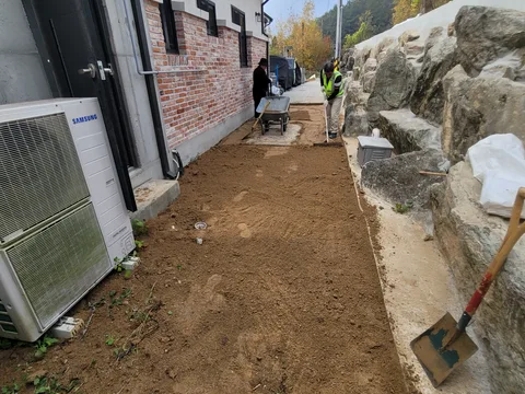 돌담을 낀 좁은 마당에서 작업자 두 명이 리어카로 자재를 옮기며 흙바닥을 정리하는 모습