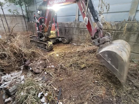 담장에 둘러싸인 좁은 마당에서 소형 굴착기로 잡초와 흙을 걷어내는 철거 작업