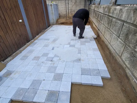 목재 담장을 따라 좁은 마당에서 작업자 한 명이 회색 블록을 배열하고 있는 모습