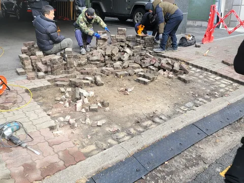 도로변 인도에서 파손된 벽돌 블록을 걷어내고 다양한 색상의 조각들을 정리하며 재시공을 준비하는 현장, 라바콘으로 작업 구간을 표시한 모습