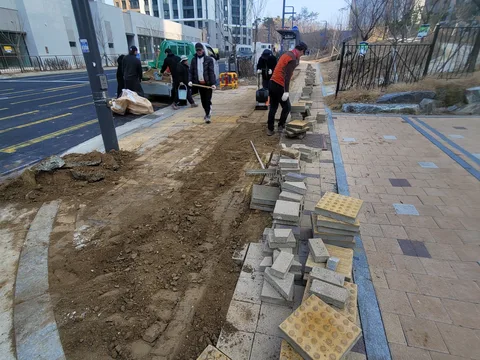 아파트 단지 담장 옆 보도에서 작업자들이 노란색 점자블록과 회색 인터로킹 블록을 함께 배열하는 시공 장면