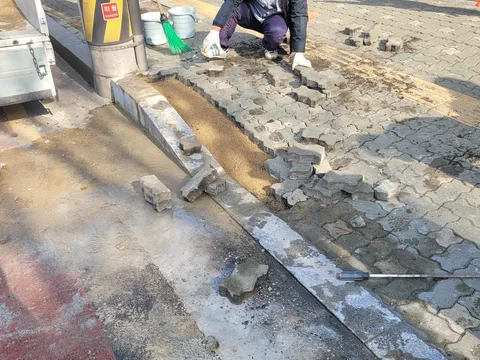 볼라드 기둥 옆 곡선 경계석 구간에 모래를 깔고 인터로킹 블록을 맞춰 배열하는 작업자